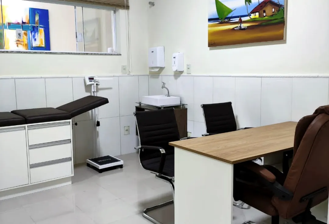 Sala de avaliação da clínica, com mesa, cadeiras e maca